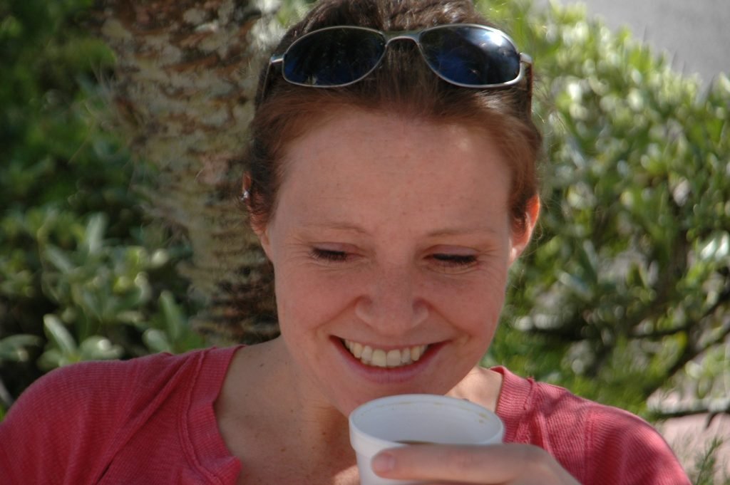 Jennifer Kraft smiling while holding a cup of coffee outdoors, wearing a casual red top and sunglasses on her head.